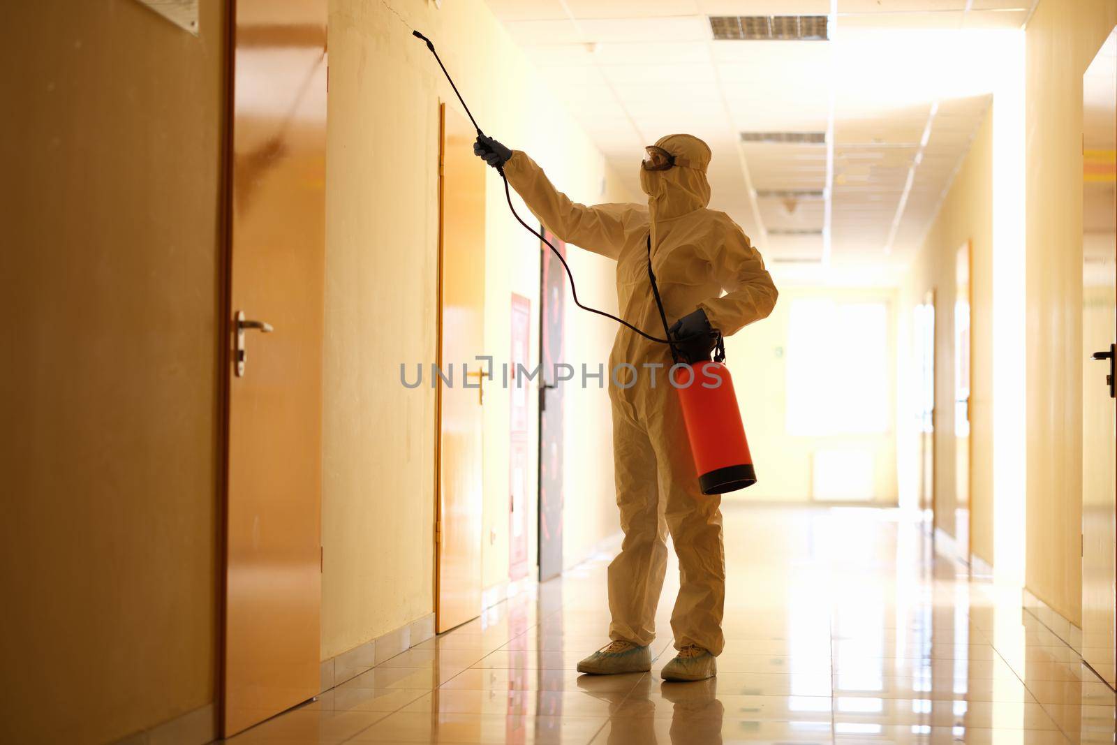 Person wearing white chemical protection suit holding spray bottle with disinfectant chemicals by kuprevich