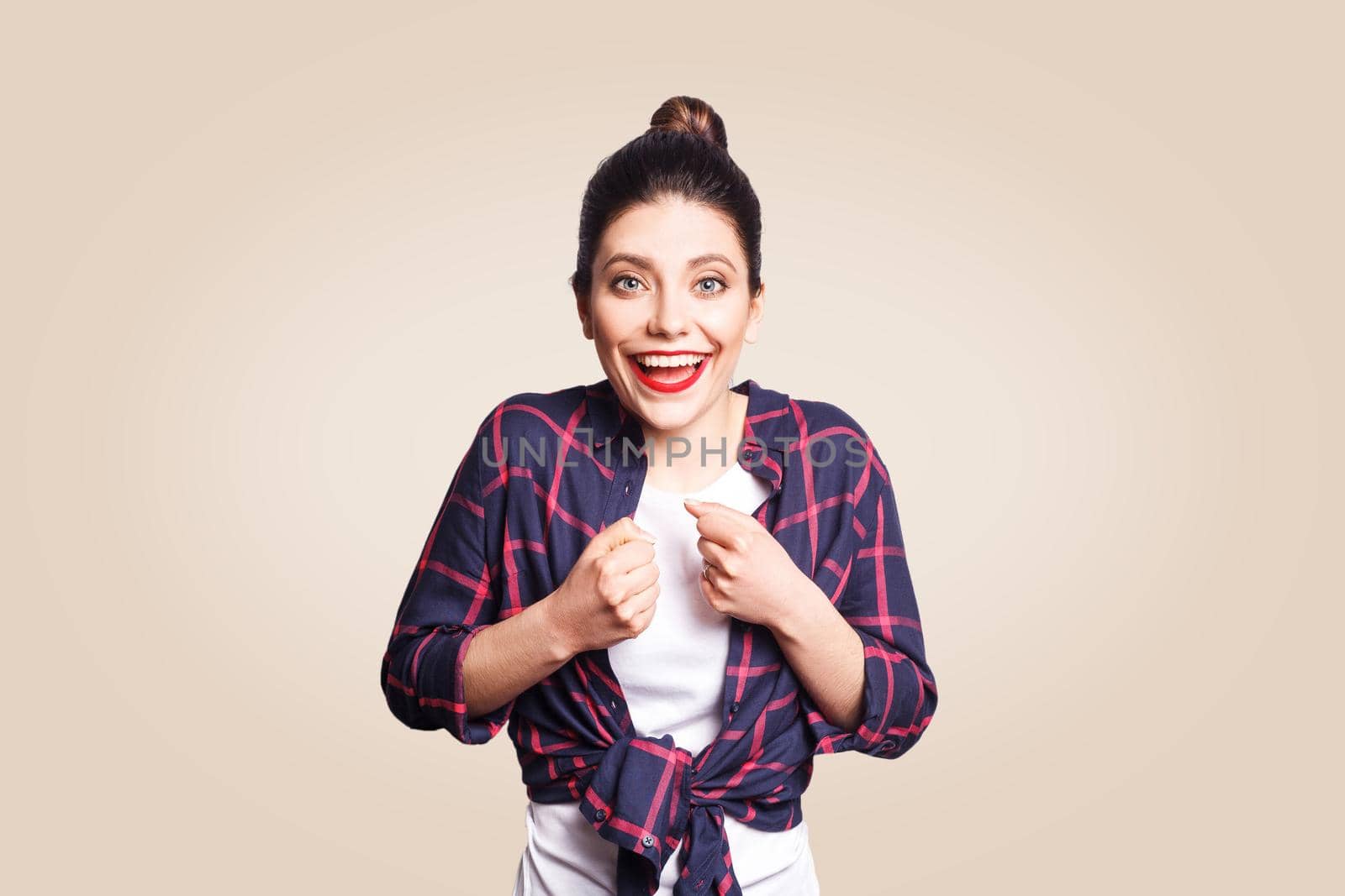 Happy excited young woman with black bun hair screaming or exclaiming, opening mouth widely, gesturing with hands, looking shocked after winning for the first time in her life. beige background.