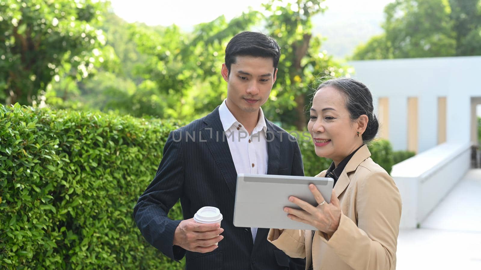 Mature businesswoman sharing idea and explaining business plan to her business partner while standing outside modern office building by prathanchorruangsak