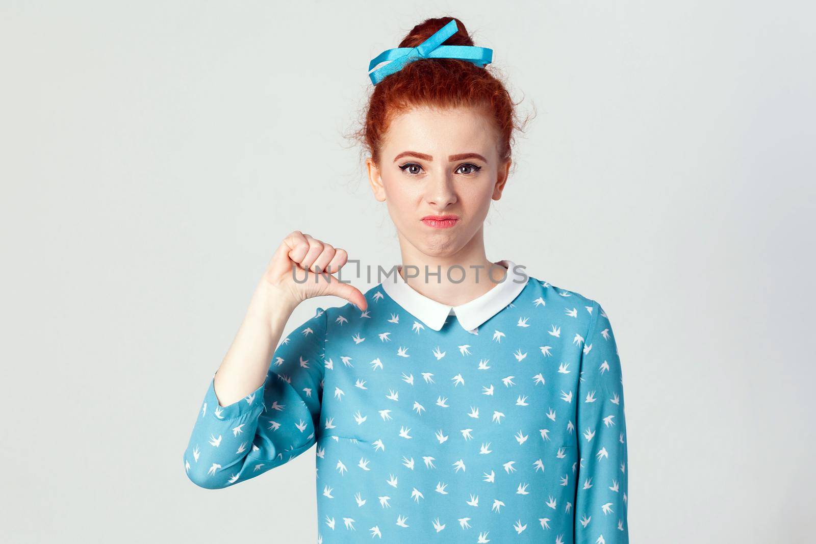 Young beautiful unhappy girl with blue dress showing thumb down. Studio shot on light gray background.