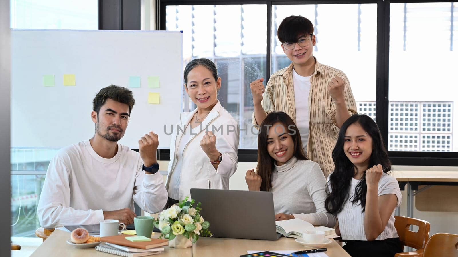 Successful mature businesswoman and team cheering with clenched fist, celebrating corporate achievement at meeting room.