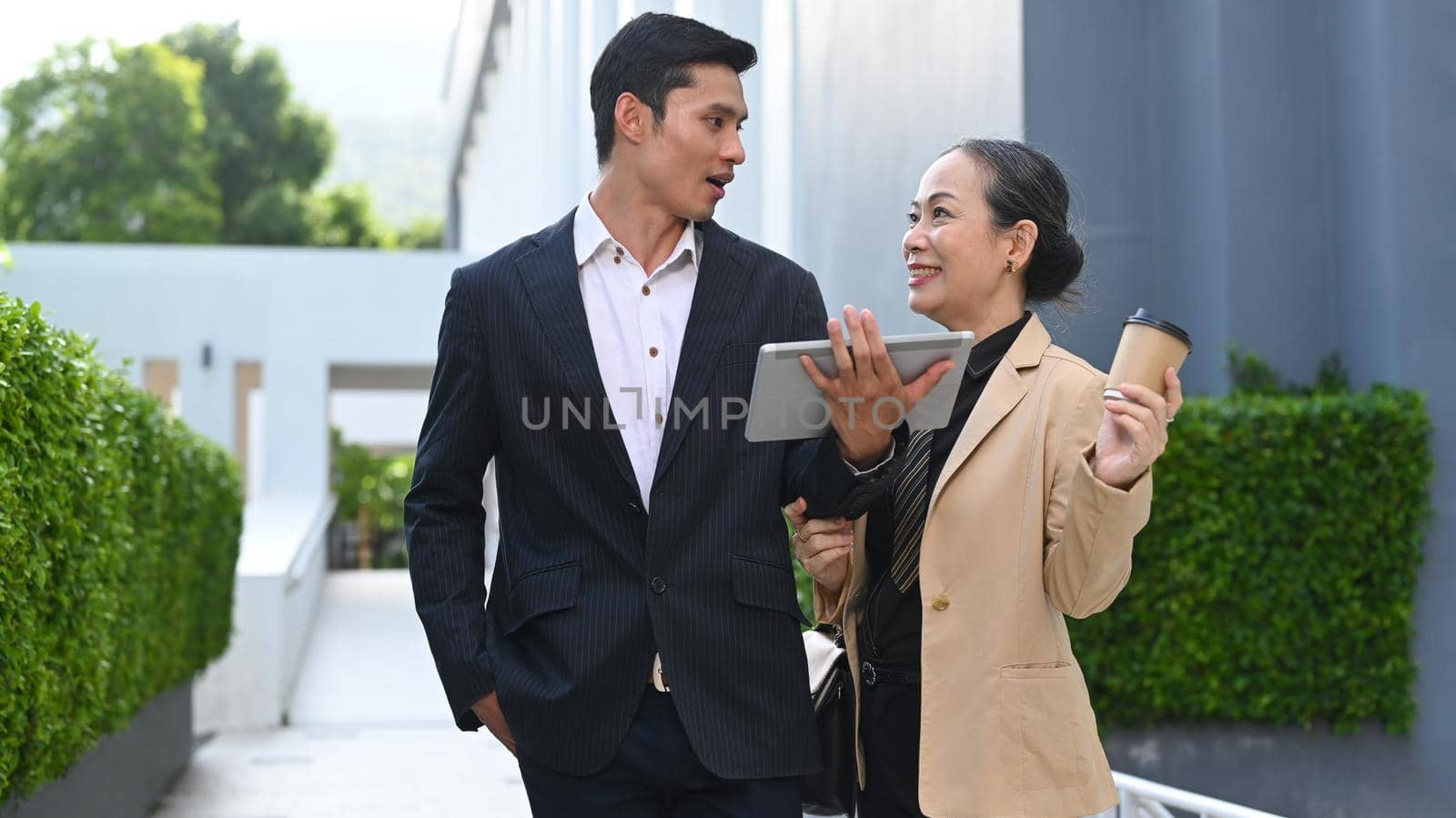 Smart young asian businessman discussing business plan with senior businesswoman while walking outside modern office building.