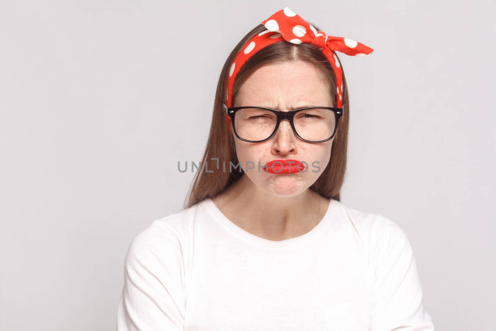 sad unhappy portrait of beautiful emotional young woman looking at camera in white t-shirt with freckles, black glasses, red lips and head band. indoor studio shot, isolated on light gray background.