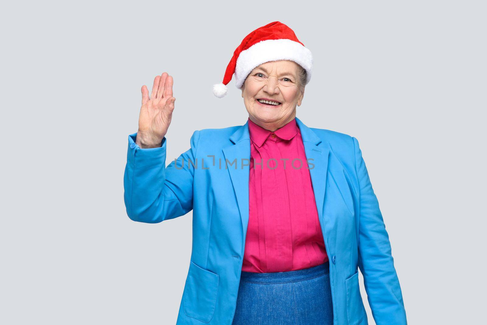 Hi, nice to see you. happy toothy smiling colorful casual style aged woman with blue suit and christmas santa red cap standing and looking at camera. indoor, studio shot, isolated on gray background