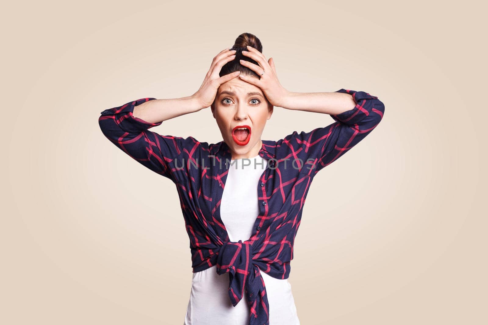 Portrait of hysterical Caucasian girl with bun hair looking in despair and panic, being late for important exam or event, not knowing what to do, hands on her head, mouth wide open. looking at camera.