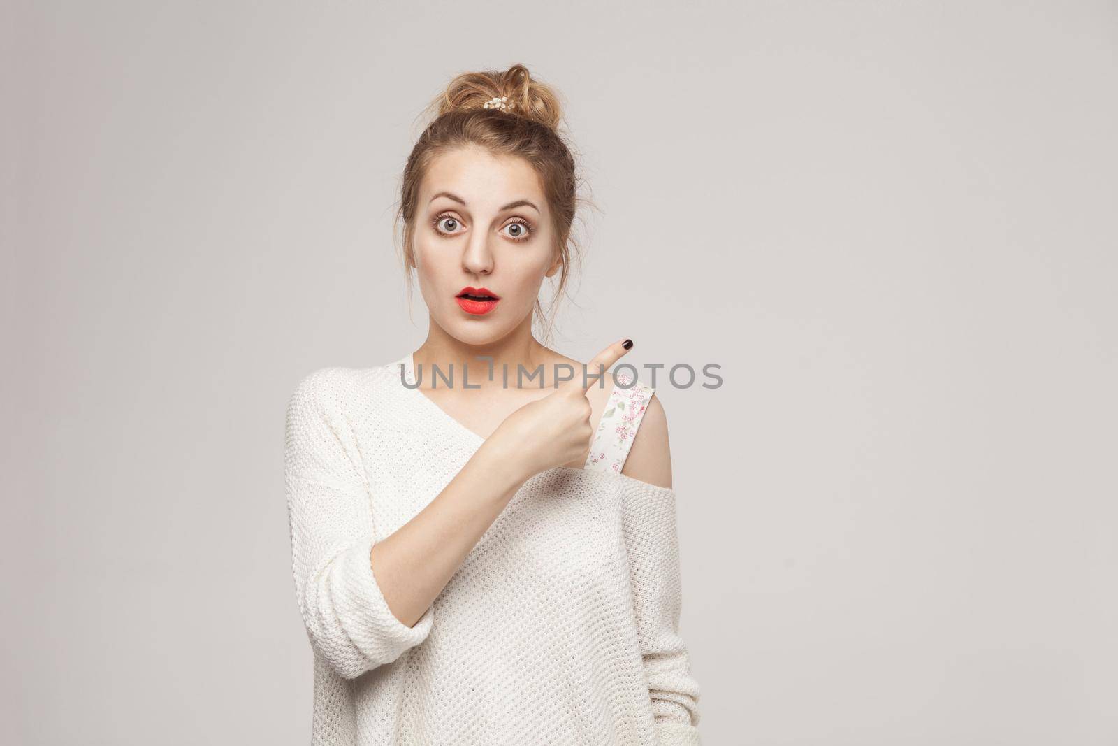 Amazement blonde woman pointing finger at copy space. Studio shot, gray wall