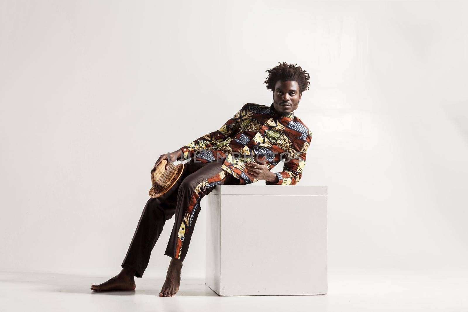 Barefooted african man uncomfortble sit on cube. Indoor, isolated on gray background