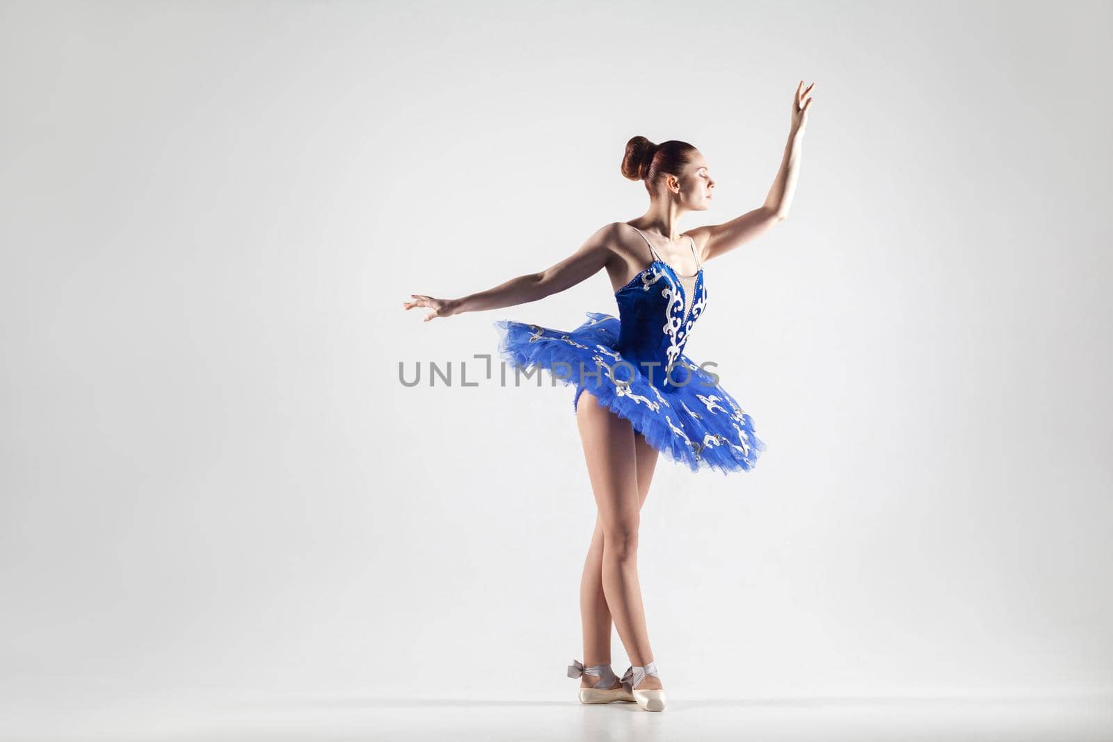 attractive ballerina with bun collected hair wearing blue dress and pointe shoes performing in white studio. indoor, studio shot.