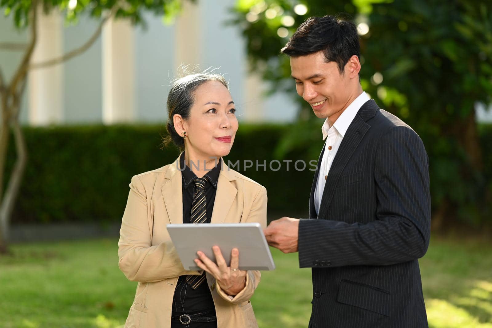 Mature female investor holding digital tablet and explaining business plan to Asian male adviser while standing at modern office district by prathanchorruangsak