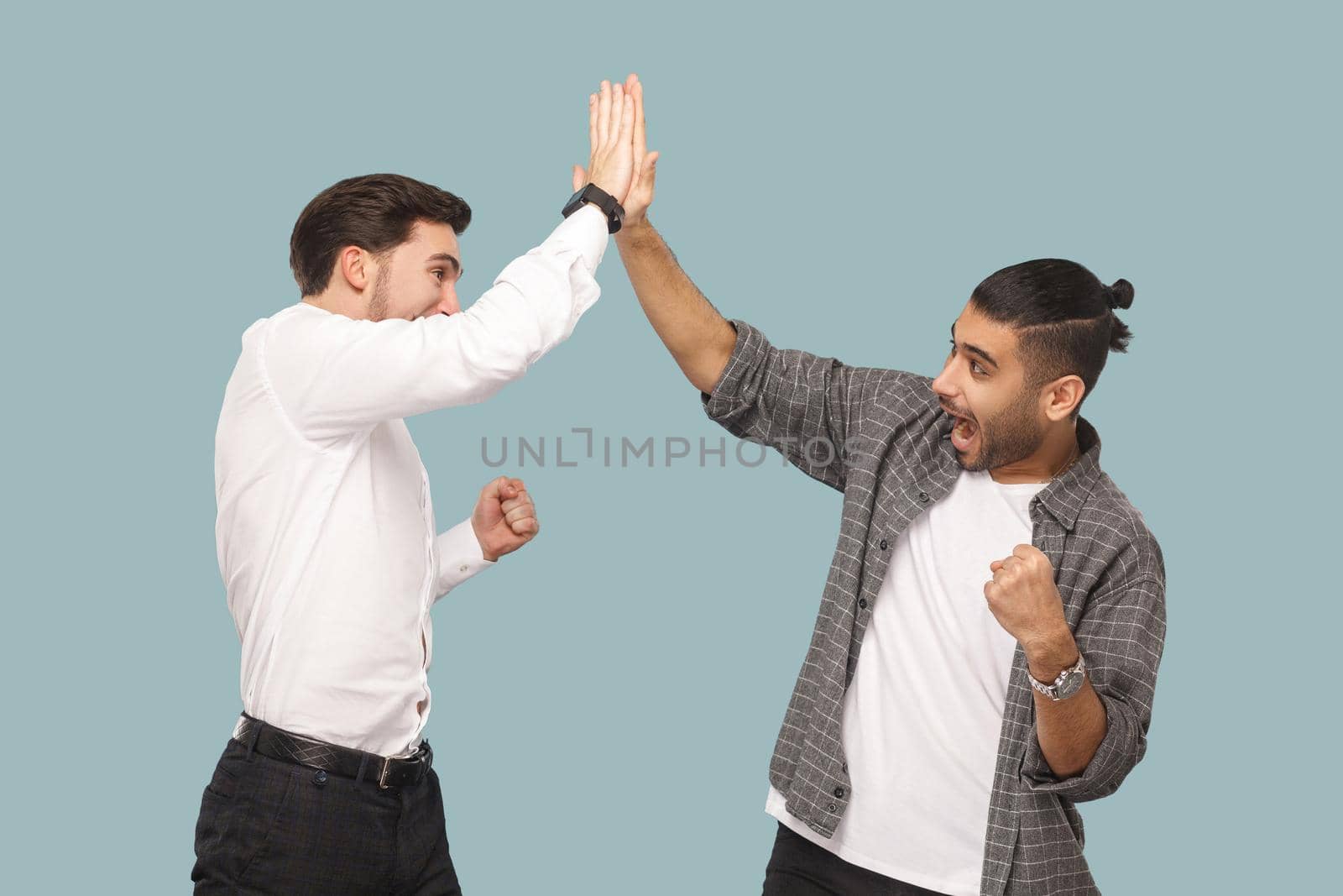 Yes we did. expression profile side view of two young handsome happy satisfied bearded partner celebrating triumph together, giving hi five hands. indoor studio shot, isolated on light blue background