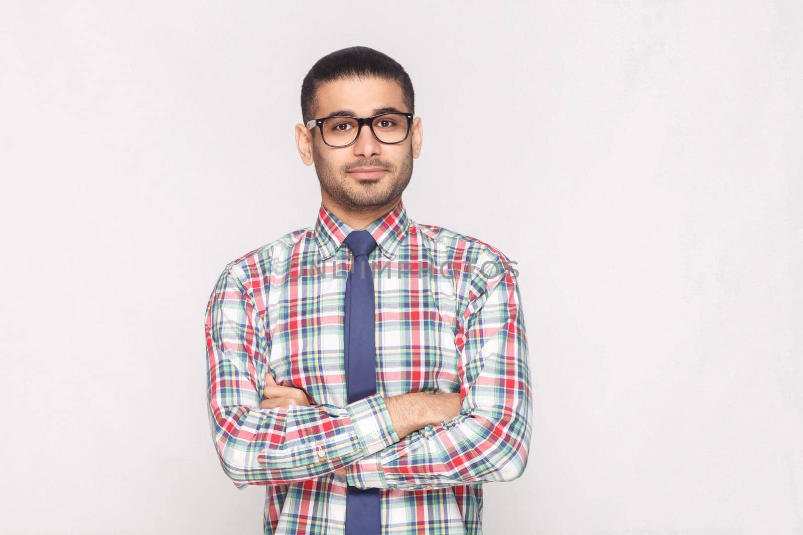 portrait of happy handsome bearded businessman in colorful checkered shirt, blue tie and black eyeglasses standing in crossed hands and smiling. indoor studio shot, isolated on light grey background.