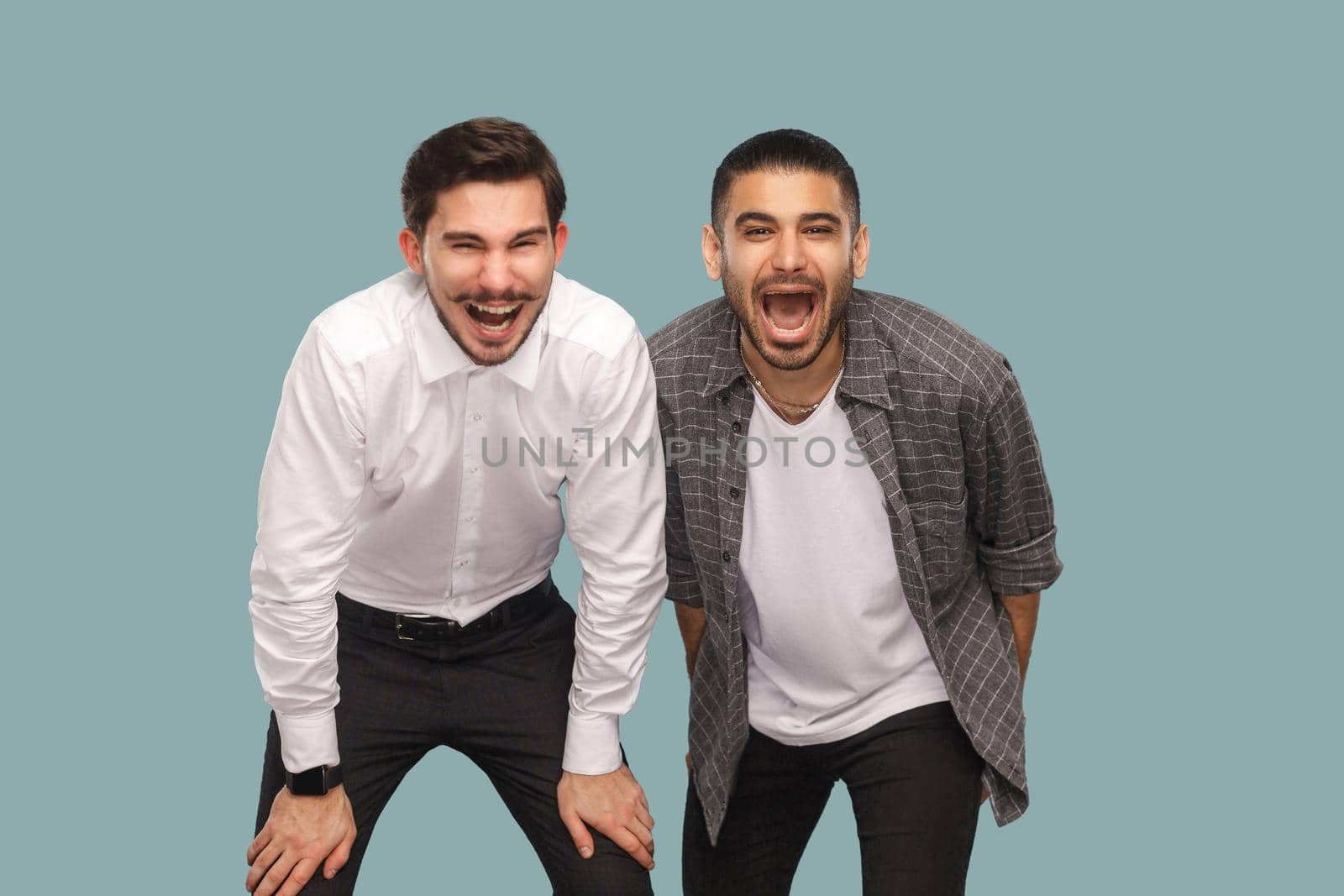 Portrait of two handsome bearded happy positive friends or partners standing and looking at camera and laughing with open mouth. indoor studio shot, isolated on light blue background.