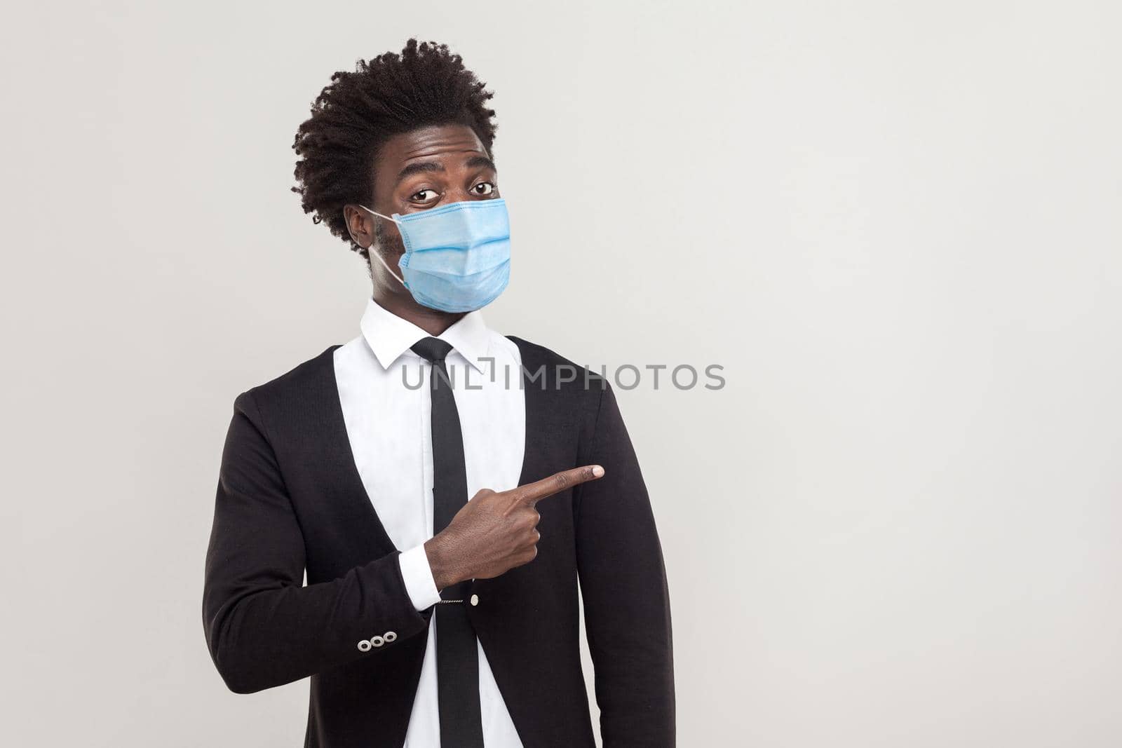 Portrait of funny young handsome worker man wearing black suit with surgical medical mask standing showing and pointing at empty background copyspace. indoor studio shot isolated on gray background.