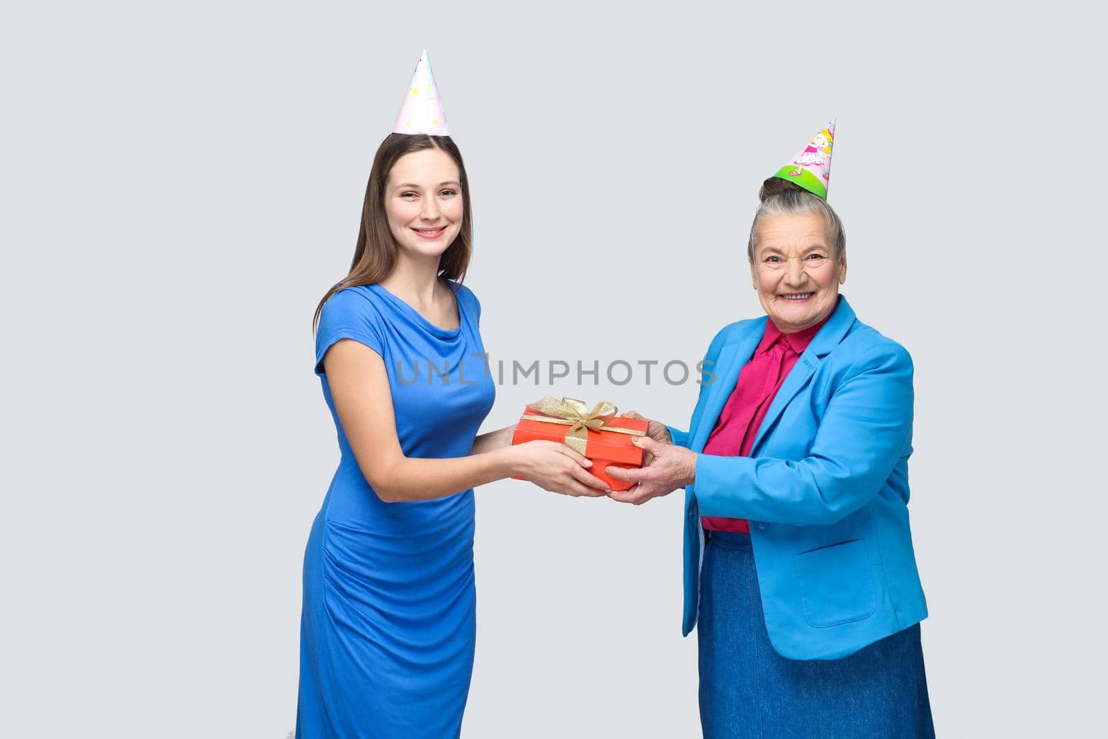 Grandmother in blue suit and hat holding red gift box and giving to her lovely granddaughter on her birthday. family happiness life event celebration. indoor studio shot, isolated on gray background