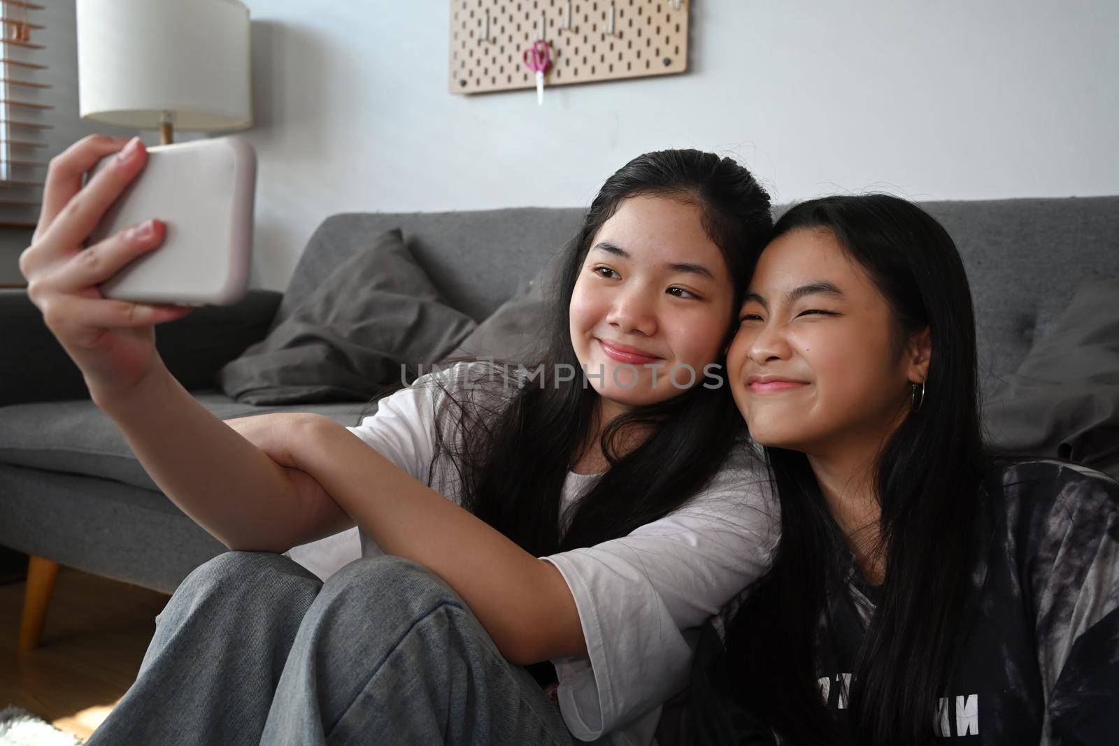 Two Asian girls taking selfie with smart smartphone while sitting together in living room. by prathanchorruangsak