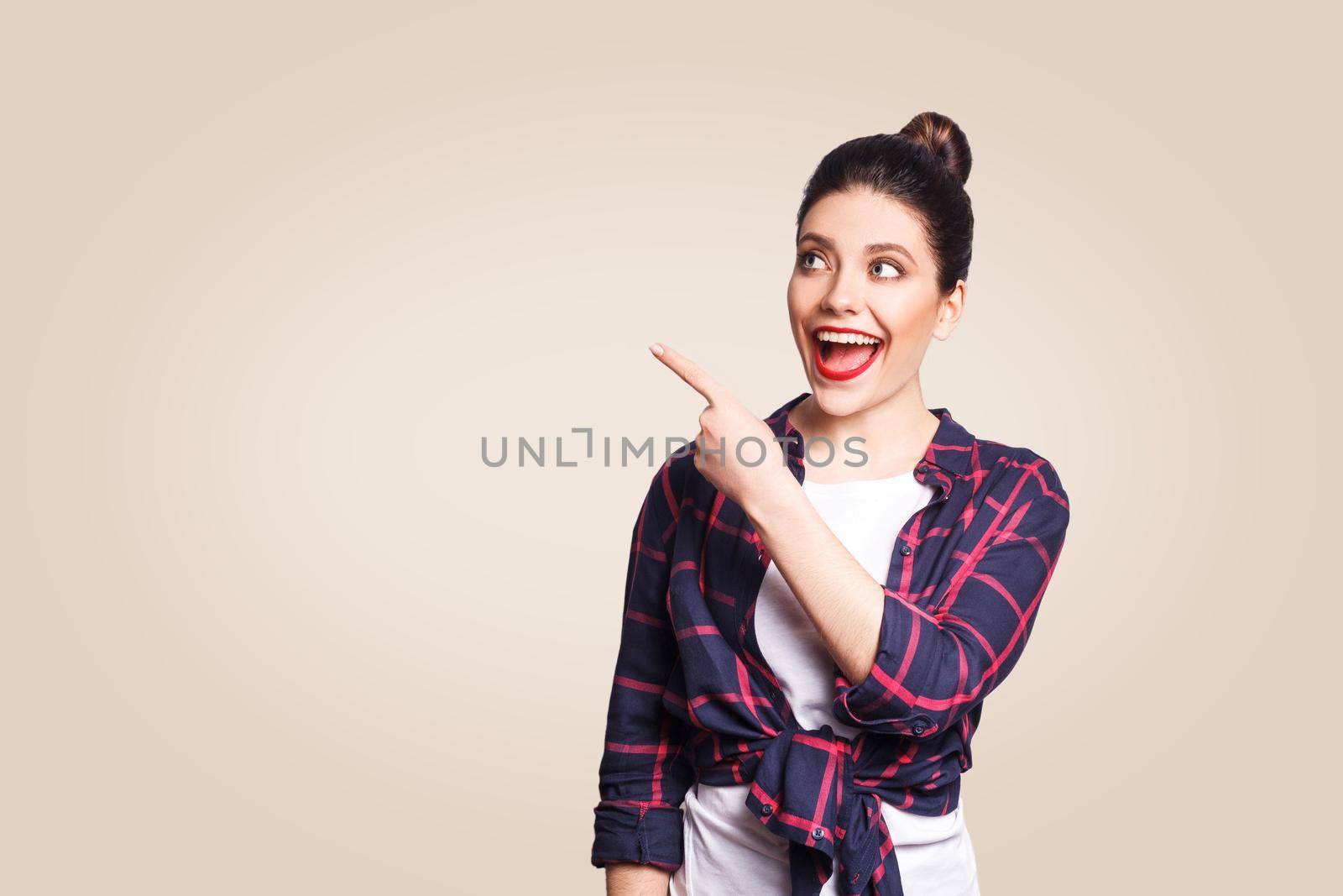 Young happy girl with casual style and bun hair pointing her finger sideways, demonstrating something on beige blank wall with copy space for your information or promotional content.