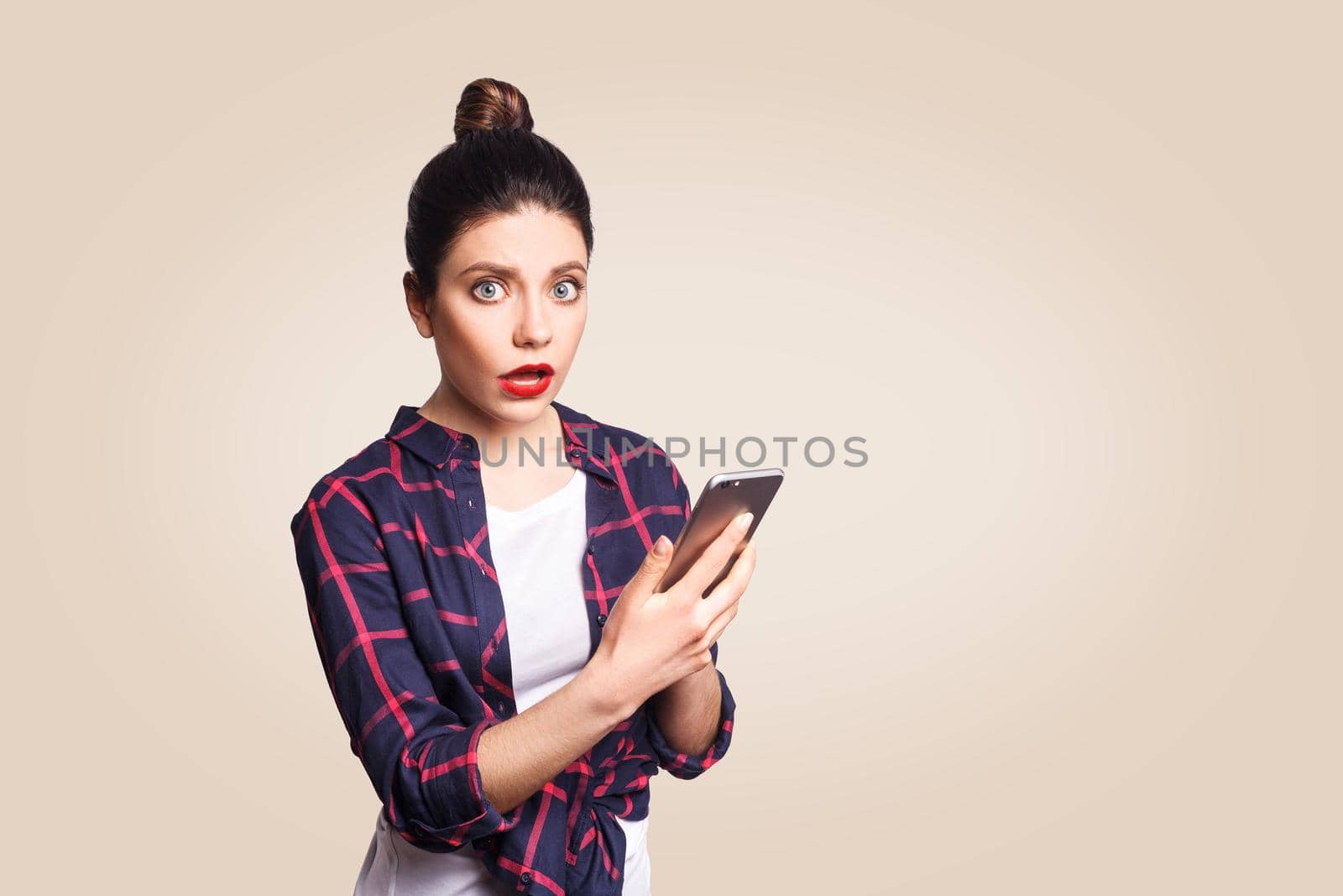 Young beautiful woman in casual style holding phone and surprised. shocked and looking at camera. studio shot on beige background.