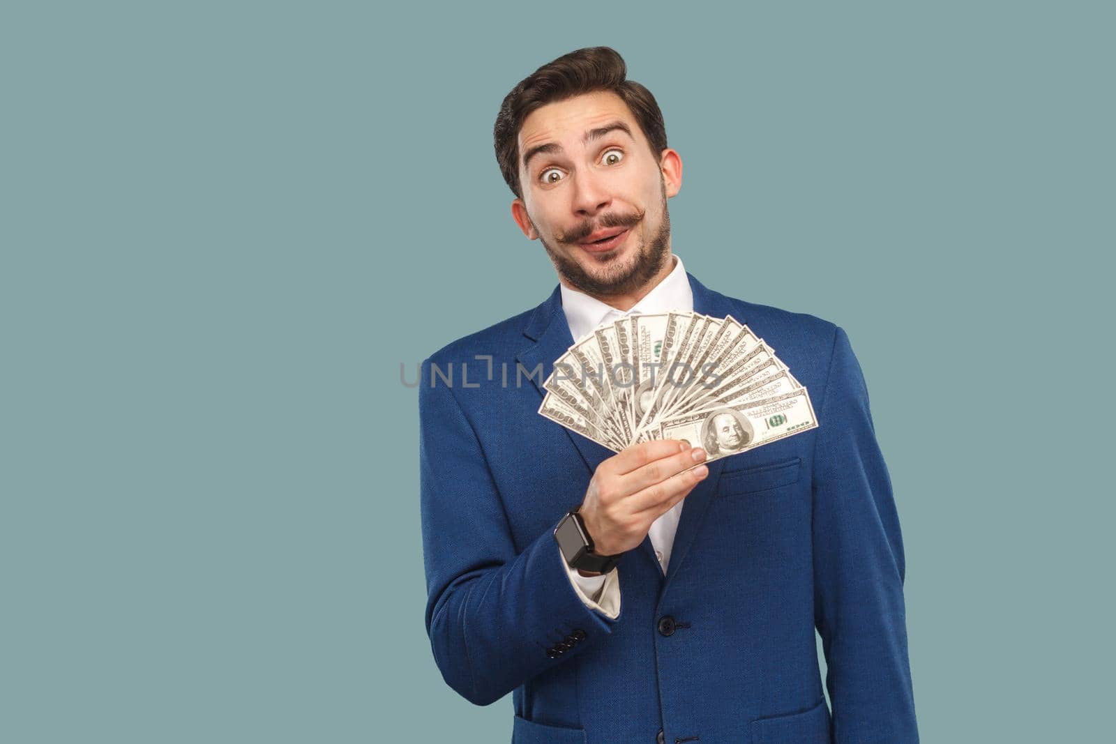 Handsome businessman in blue jacket standing and holding many dollars in hand and looking at camera with amazed surprised face and big eyes. Indoor, studio shot isolated on light blue background.