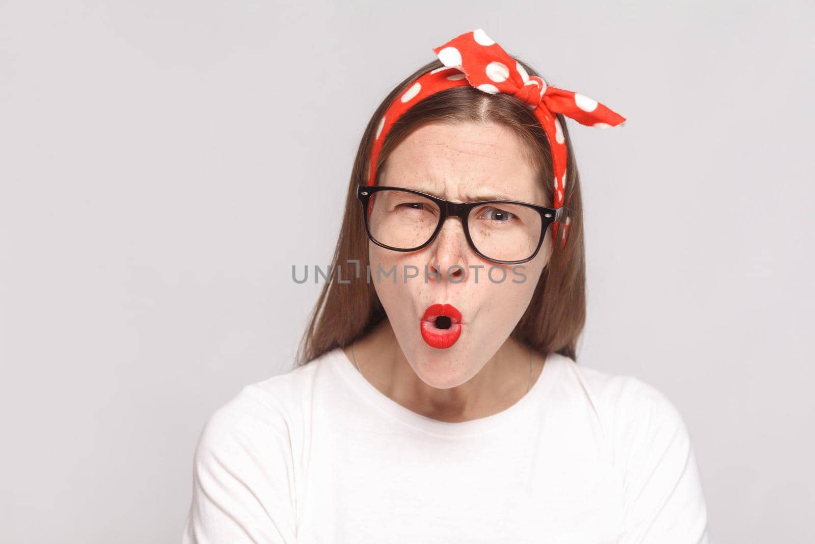 what? shocked face closeup portrait of beautiful emotional young woman in white t-shirt with freckles, black glasses, red lips and head band. indoor studio shot, isolated on light gray background.