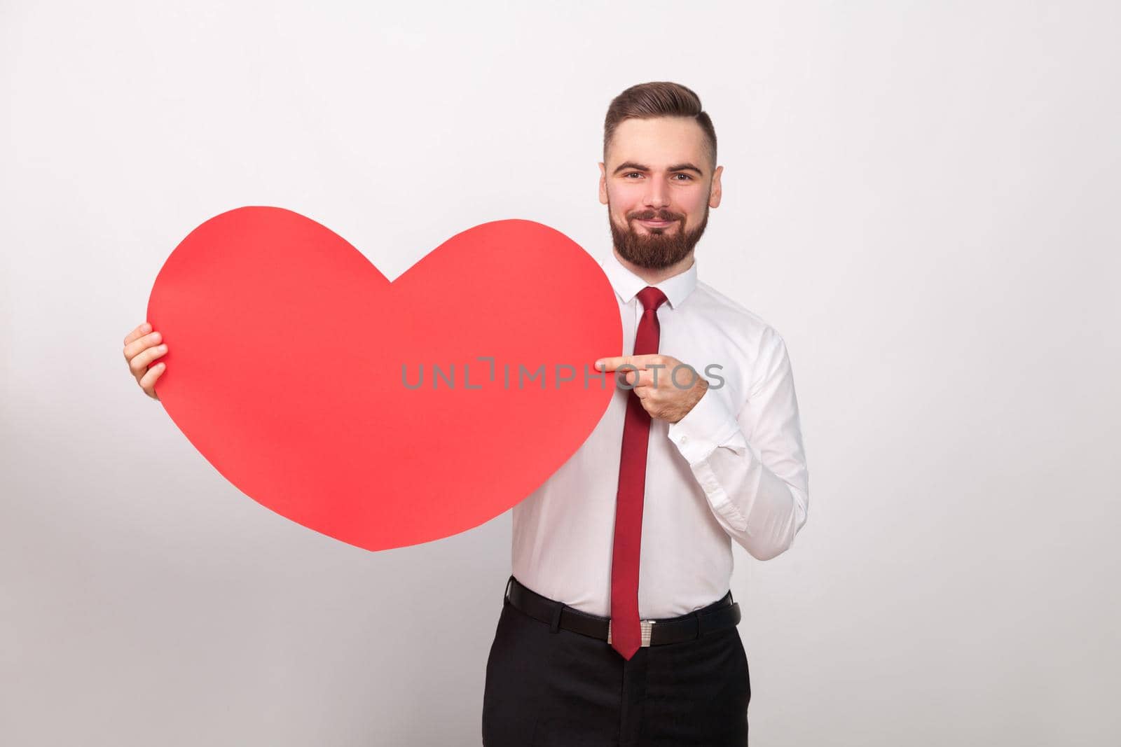 Perfect businessman smiling, pointing finger at big red heart. Indoor, studio shot, isolated on gray background