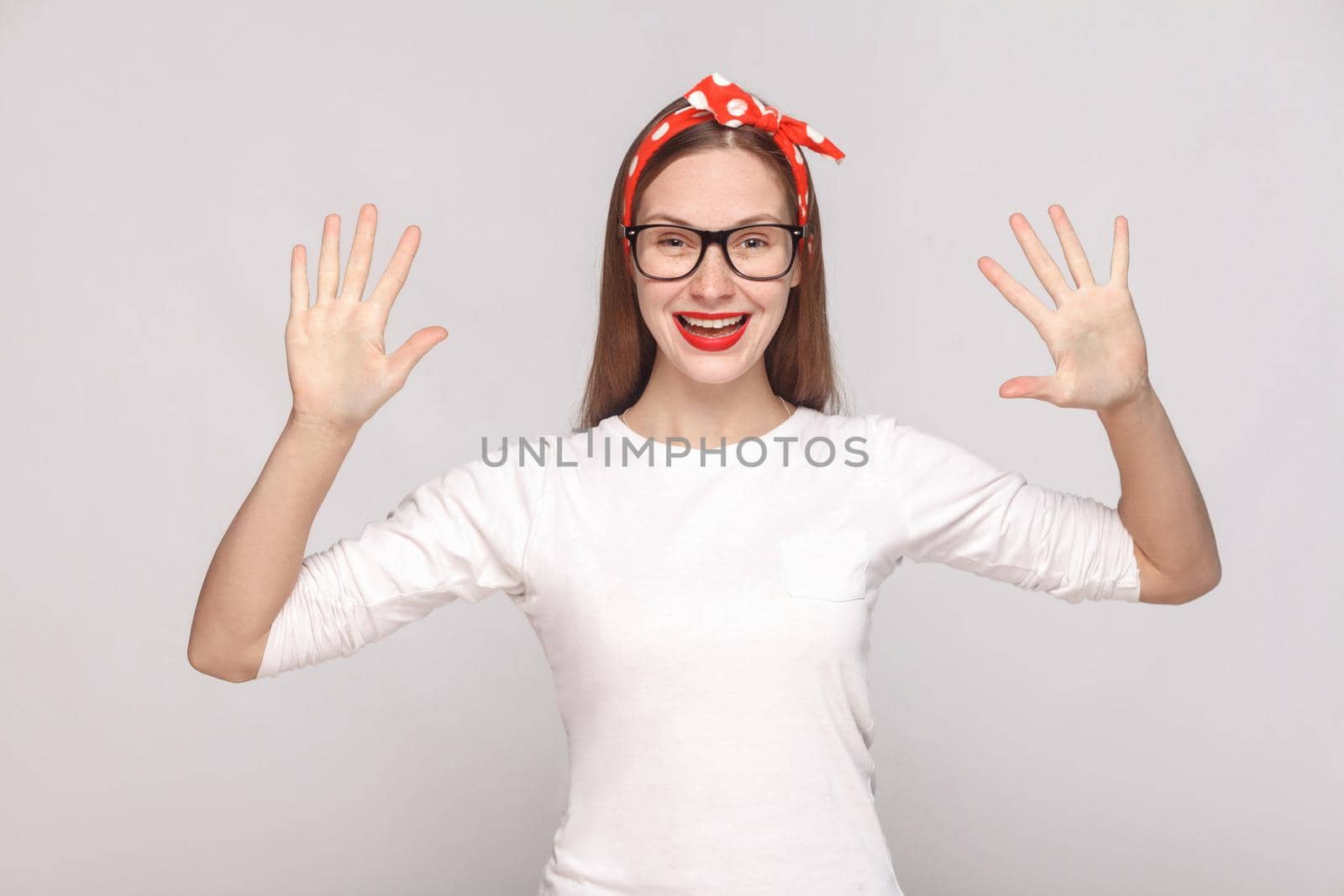 hey, nice to see you. portrait of beautiful emotional young woman in white t-shirt with freckles, black glasses, red lips and head band. indoor studio shot, isolated on light gray background.