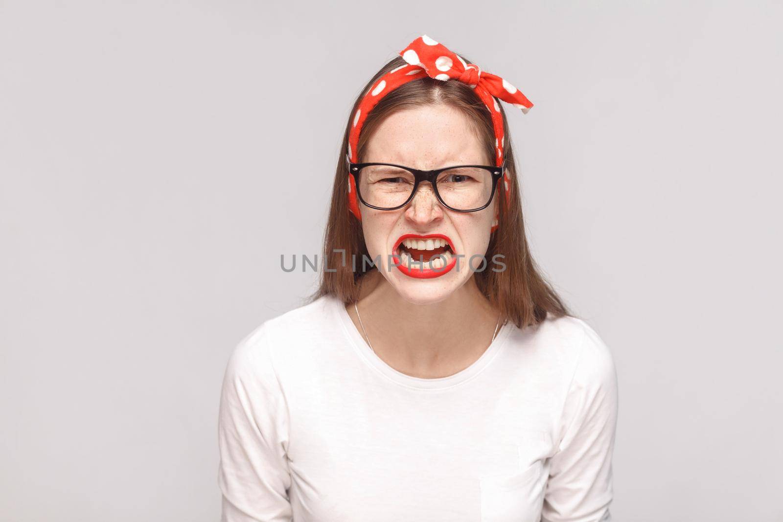 angry face screaming portrait of anger crazy bossy emotional young woman in white t-shirt with freckles, black glasses, red lips and head band. indoor studio shot, isolated on light gray background.