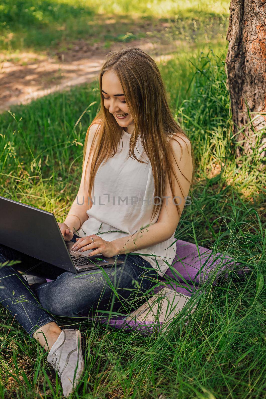 On the banner, a young girl works with a laptop in the fresh air in the park, sitting on the lawn. The concept of remote work. Work as a freelancer. The girl takes courses on a laptop and smiles