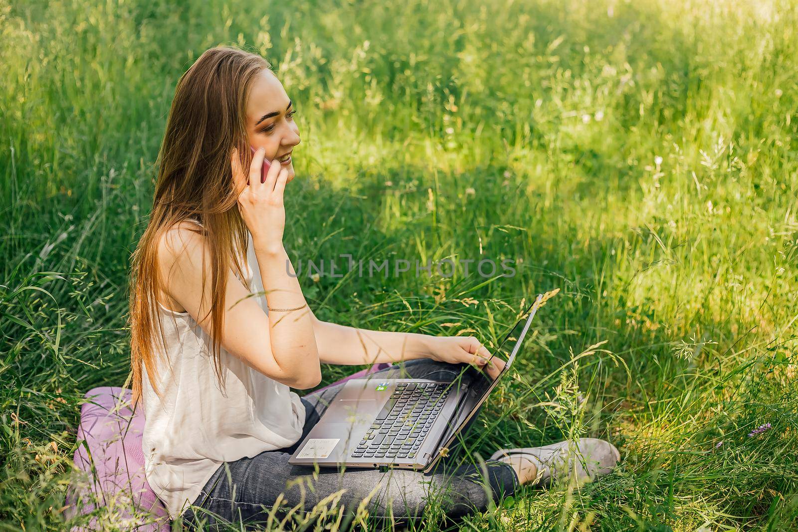 girl sits on the grass and works at a laptop. talking on the phone. freelance. selfeducation. the concept of remote learning and outdoor work. by Anyatachka