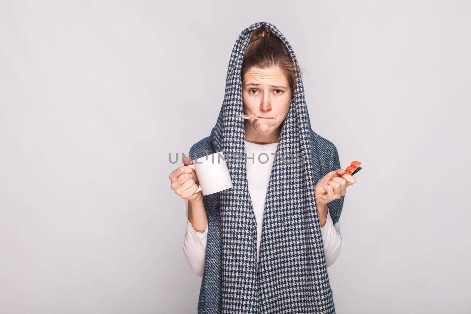 Young woman with gray scarf, holding cup, thermometer and pills. Indoor shot.