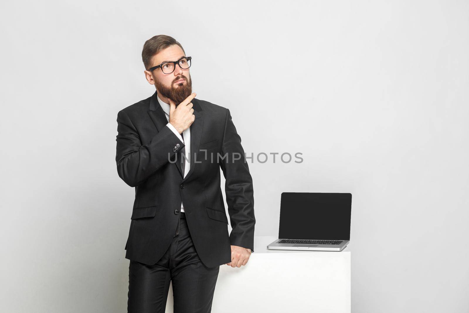 Serious confident thoughtful bearded young businessman in black suit are standing near his working place and holding his beard with concentration face. Isolated, studio shot, indoor, gray background