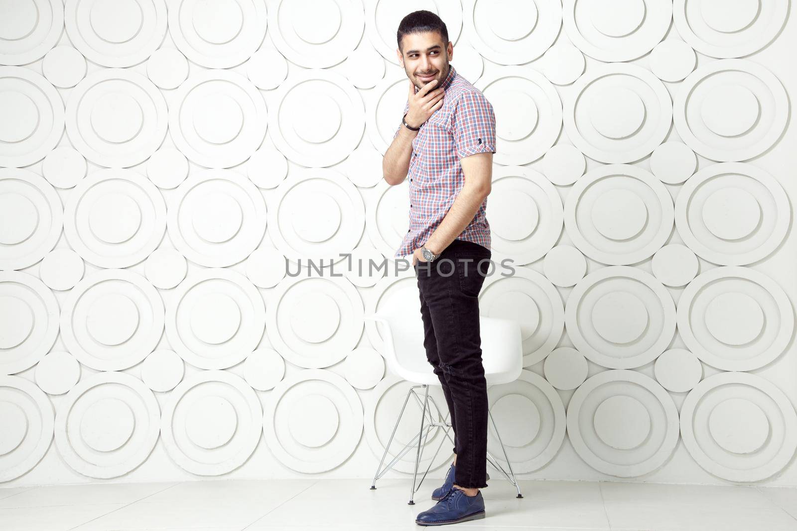 Young bearded fashion model in casual style is posing near white circle wall background. studio shot. smiling and looking away with hand on packet.