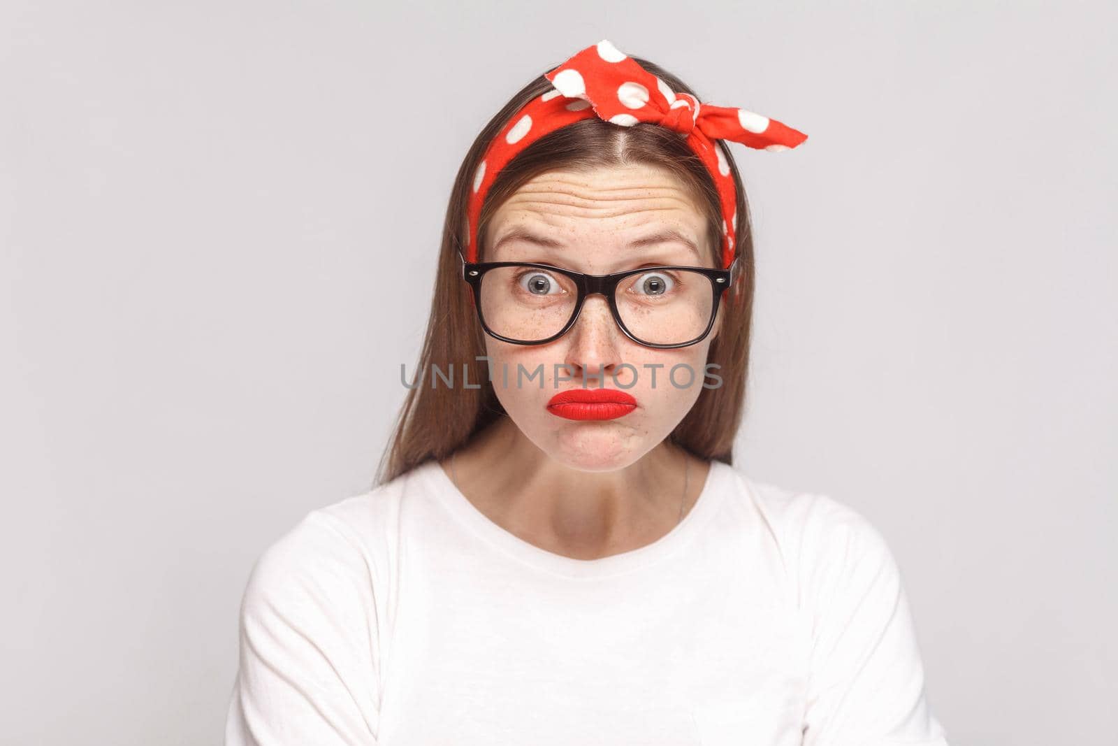 big eyes shocked face of beautiful emotional young woman in white t-shirt with freckles, black glasses, red lips and head band looking at camera. indoor studio shot, isolated on light gray background.