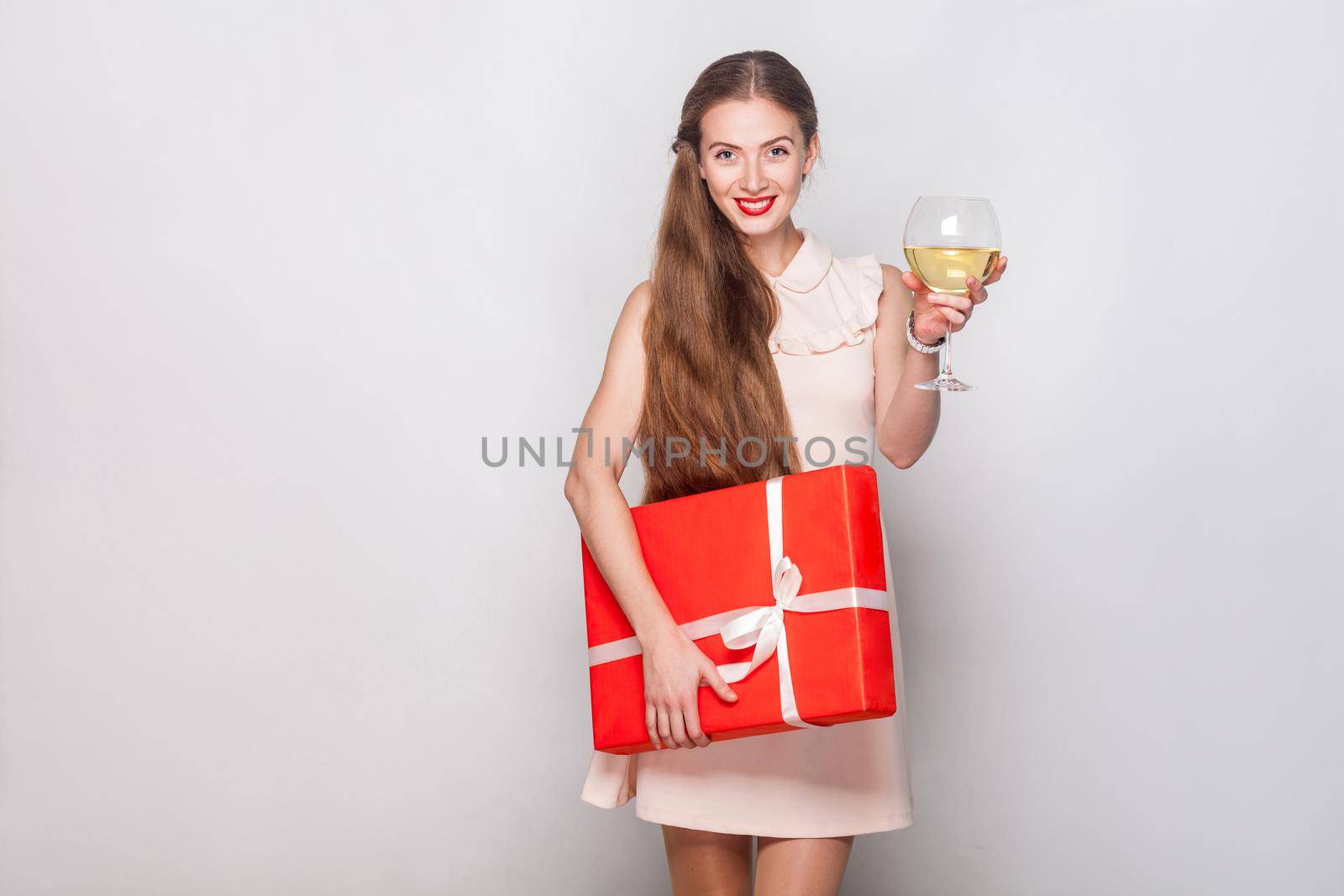 Happiness blonde woman in red cap holding champagne glass and gift box, looking at camera and toothy smiling. Studio shot. Gray background