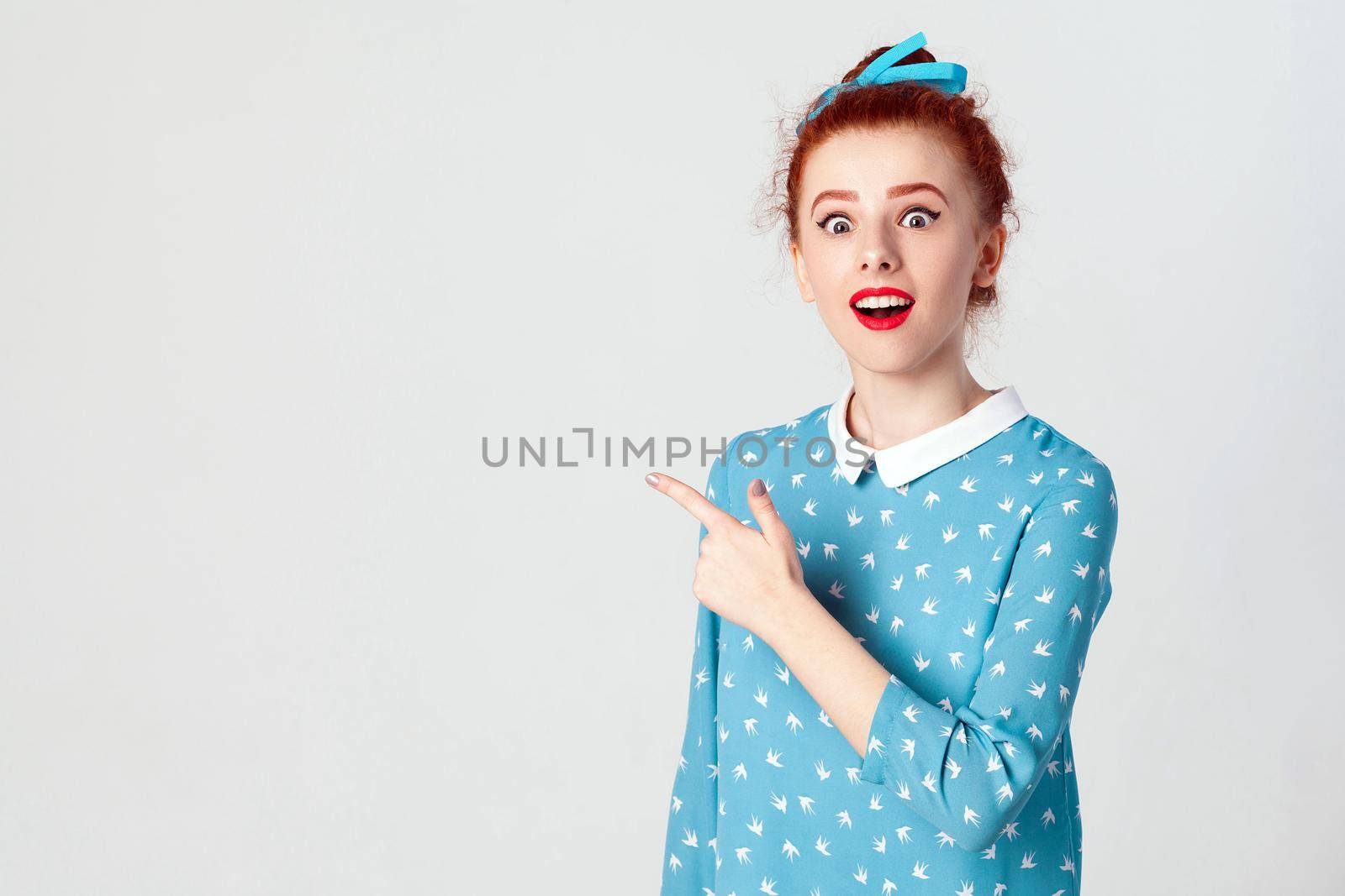 Excited young redhead caucasian girl with hair knot pointing her index finger sideways, raising eyebrows and keeping mouth wide opened, showing something surprising on grey copy space wall.