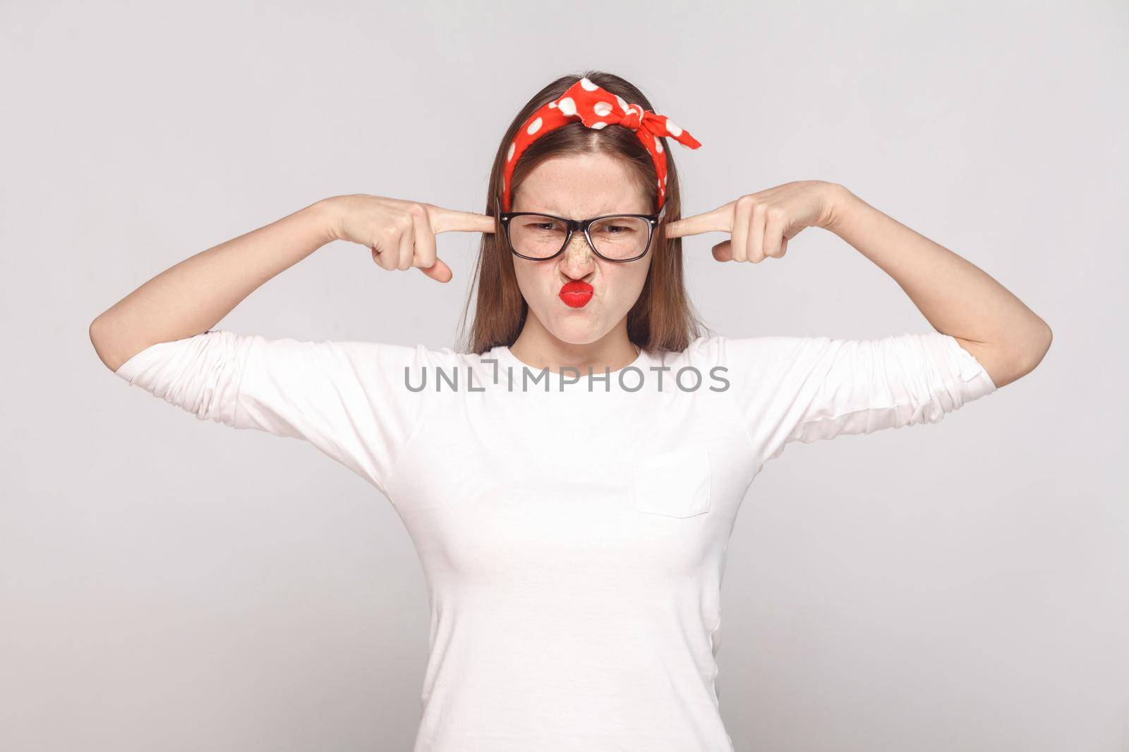 i dont want to hear you. portrait of unsatisfied emotional young woman in white t-shirt with freckles, black glasses, red lips and head band. indoor studio shot, isolated on light gray background.