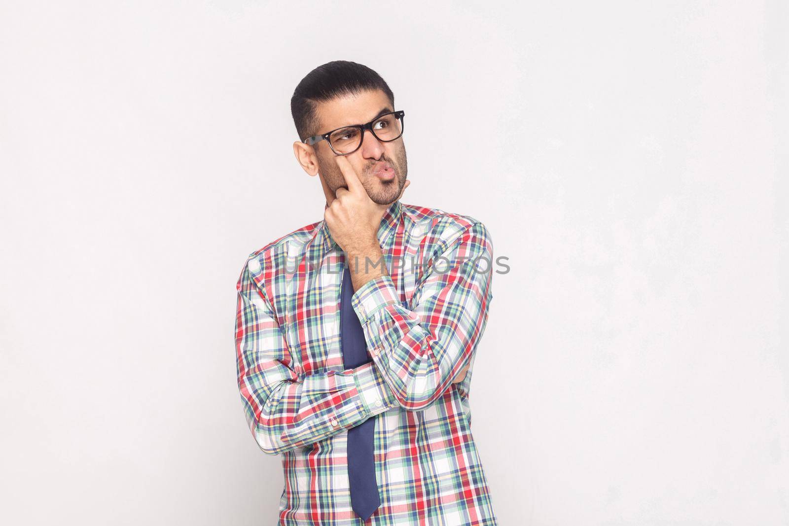 Portrait of confused handsome bearded businessman in colorful checkered shirt, blue tie and eyeglasses standing touching face and looking away. indoor studio shot, isolated on light grey background.