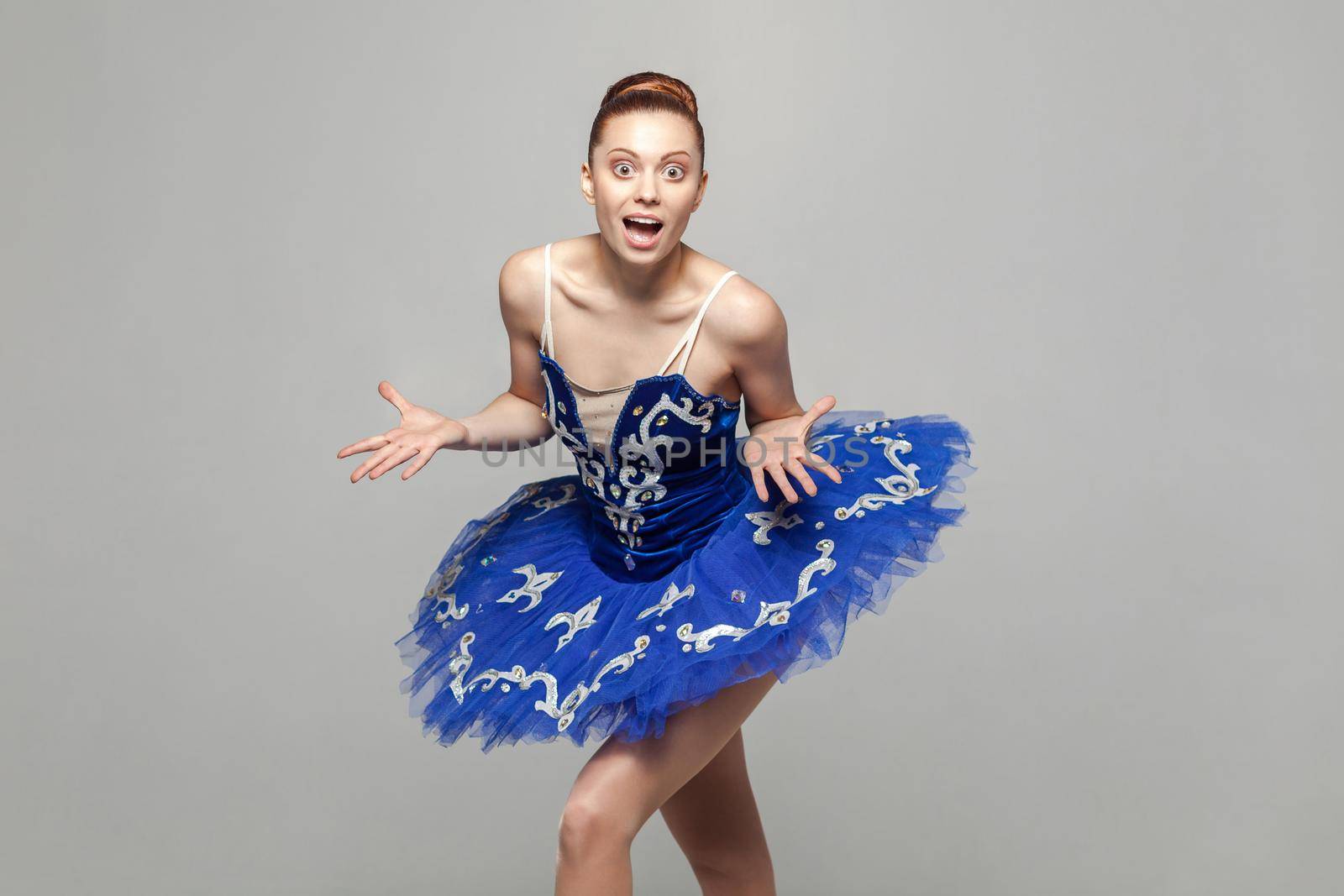 wow, its unbelievable. ballerina woman in blue costume with makeup and bun collected hair style standing against gray background looking at camera. emotion and expression concept. indoor studio shot.