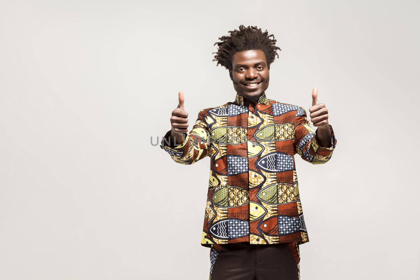 African man in traditional clothes toothy smile, showing thumbs up, loke sign. Indoor, isolated on gray background