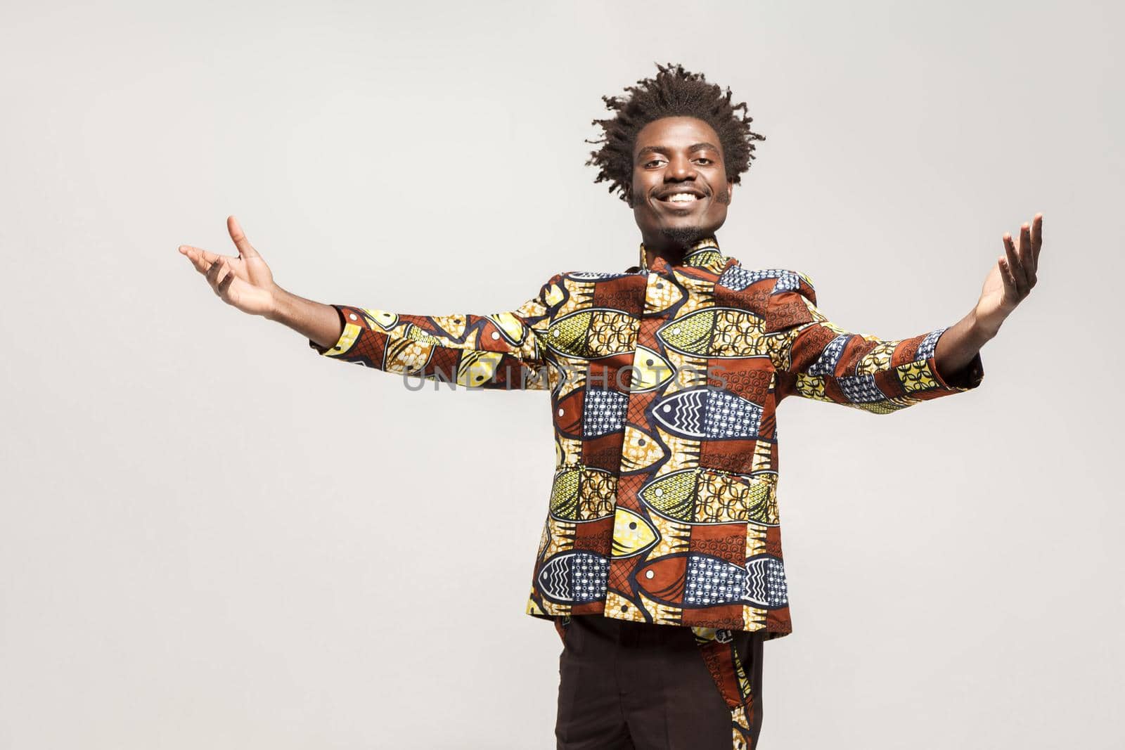 Friendly afro man in traditional clothes toothy smile, say welcome. Indoor, isolated on gray background