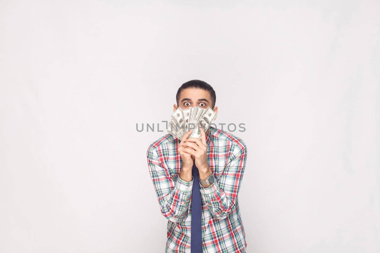 Portrait of rich handsome young adult businessman in colorful checkered shirt with blue tie standing and showing fan of dollars with big crazy eyes. Indoor, studio shot, isolated on grey background.