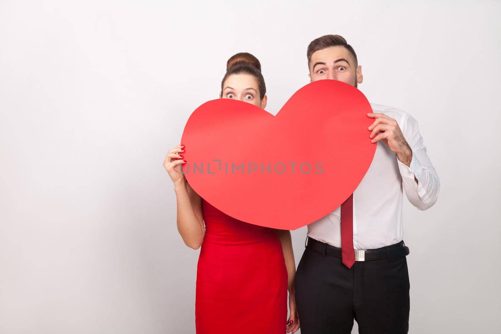 Funny couple hide and seek behind big red heart. Indoor, studio shot, isolated on gray background