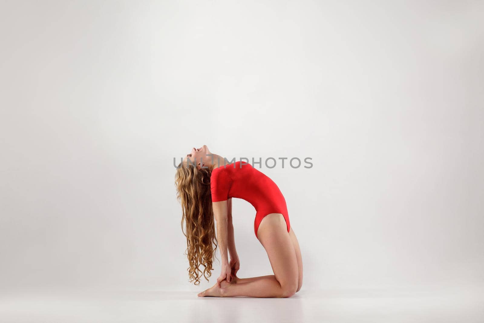 blonde woman with long curly hair in red leotard balancing on hands and knees while doing ustrasana camel yoga pose. indoor, on light gray background. healthy lifestyle and leisure activity concept