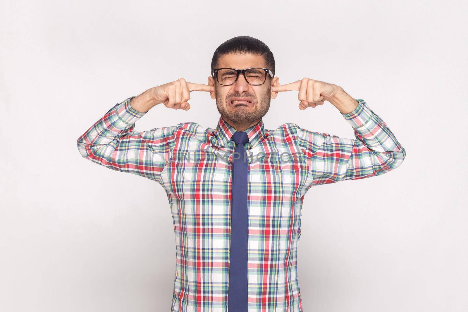 I don't want to hear. closed eyes unhappy bearded businessman in colorful checkered shirt, blue tie and eyeglasses standing with finger in ears. indoor studio shot, isolated on light grey background.