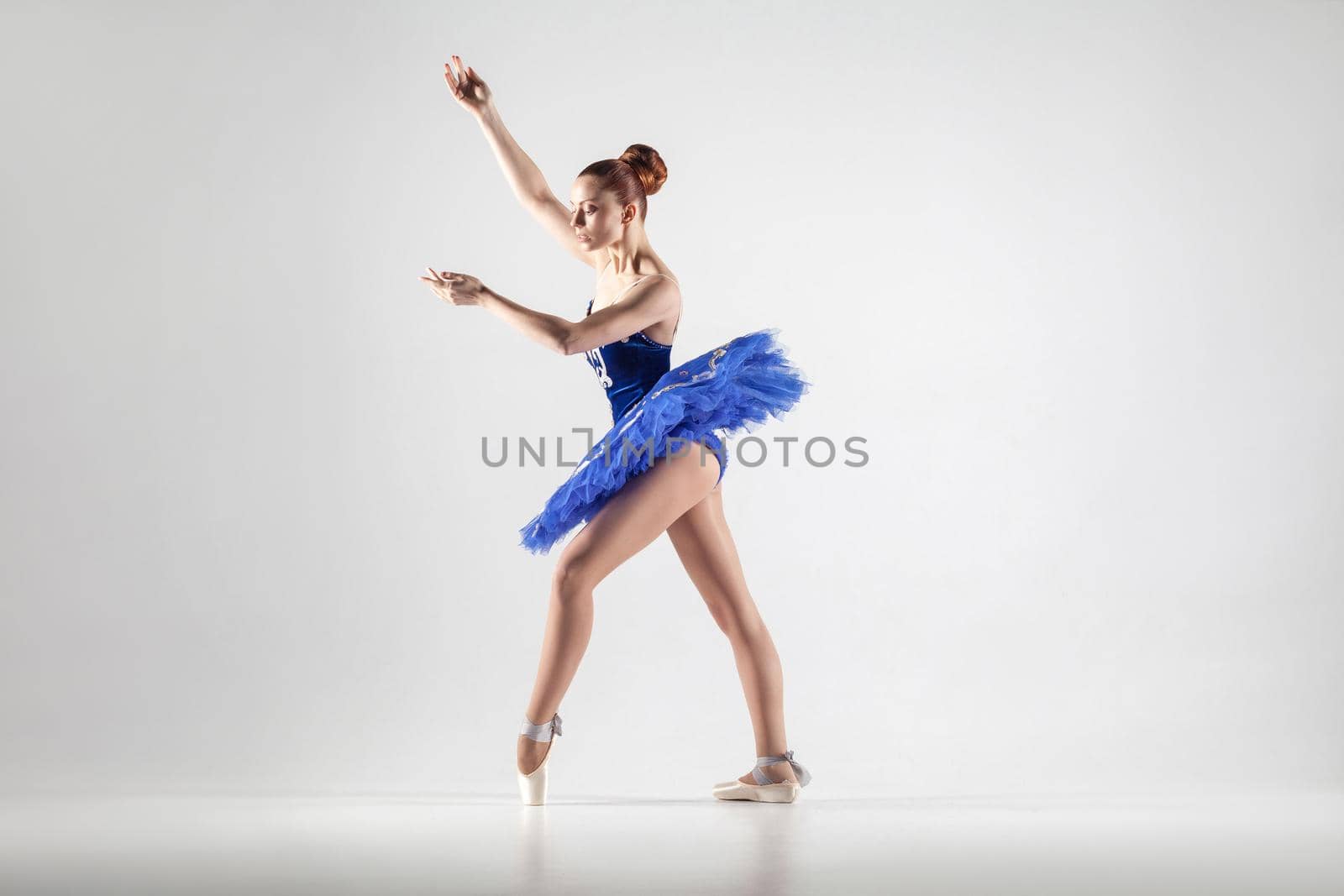 Young beautiful ballerina with bun collected hair wearing blue dress and pointe shoes dancing gracefully isolated on white background. indoor, studio shot.