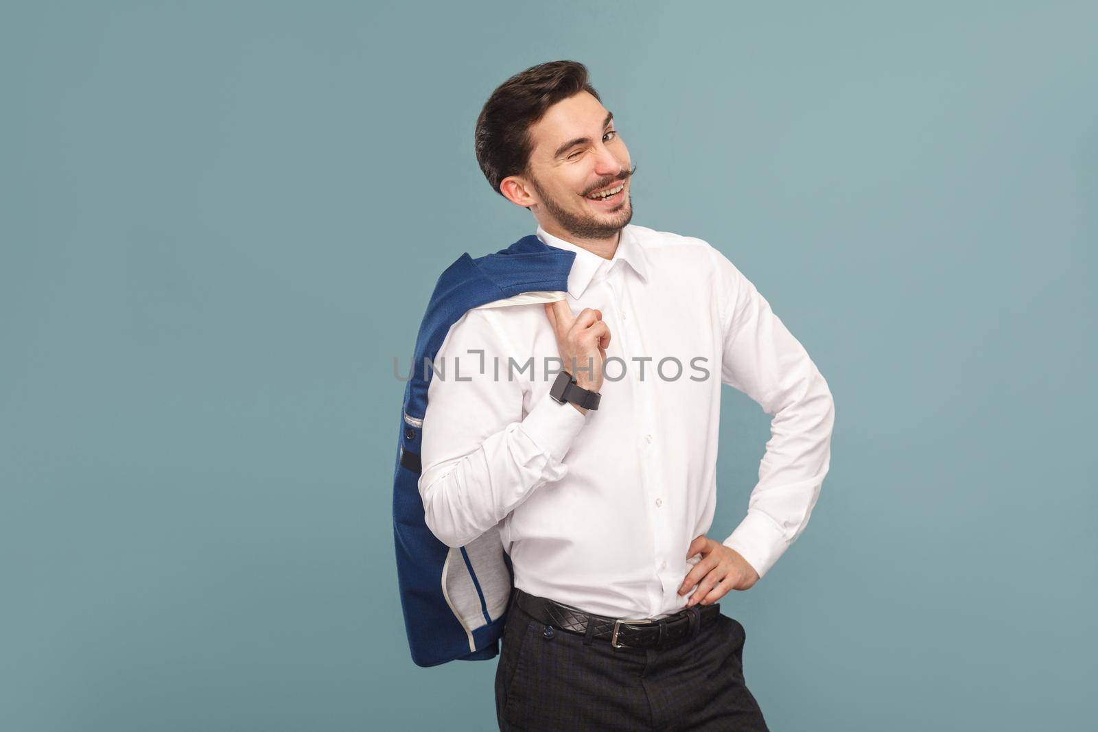 Portrait of successful boss, toothy smiling and wink of handsome bearded businessman in white shirt holding his blue jacket, with smart watch. Indoor studio shot, isolated on light blue background