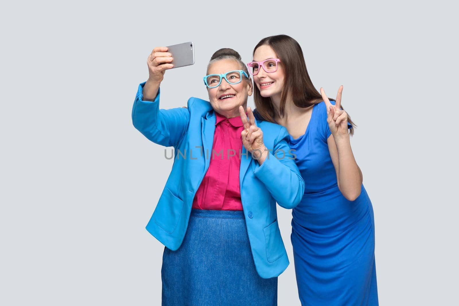 happy grandmother with granddaughter in blue dress or suit toothy smiling, standing, posing and making selfie and peace sign. Relations in the family. indoor, studio shot, isolated on gray background