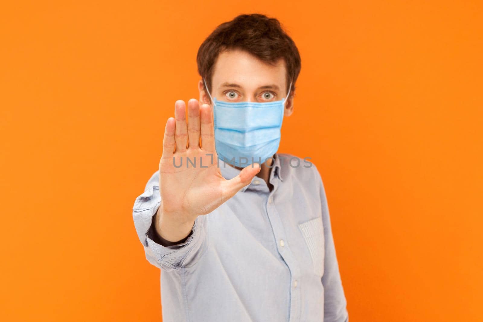 Stop! keep your distance. Portrait of angry or aggressive young worker man with surgical medical mask standing with stop hand and looking at camera. indoor studio shot isolated on orange background.