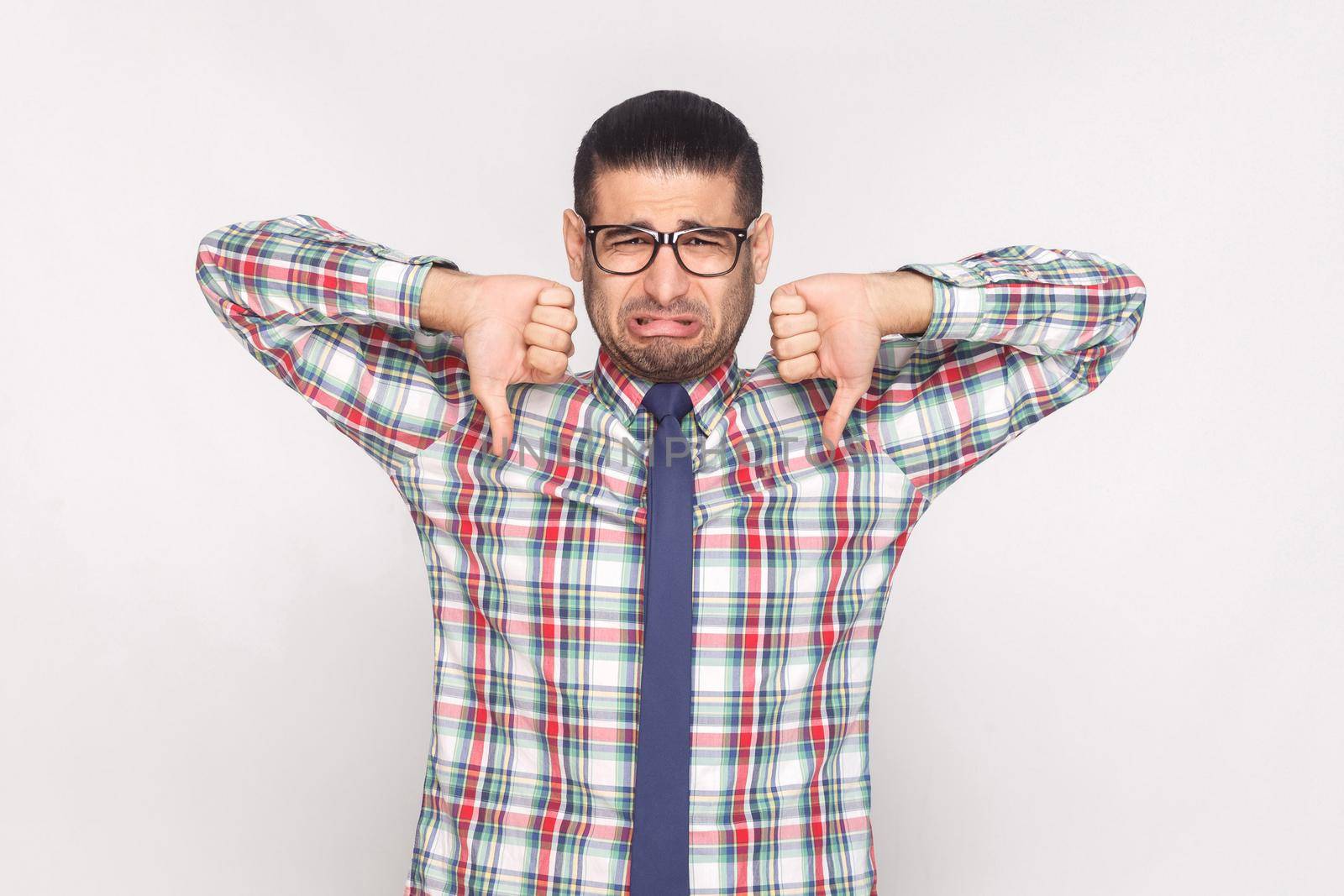 Sad unhappy bearded businessman in colorful checkered shirt, blue tie and eyeglasses standing and looking with thumbs up and dislike gesture. indoor studio shot, isolated on light grey background.