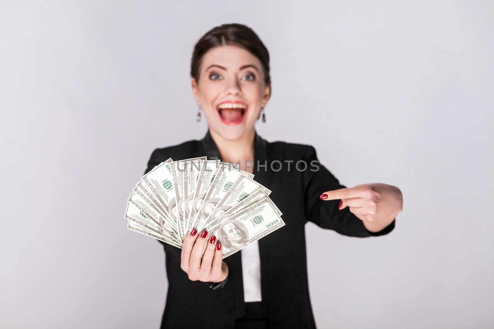 Well dressed woman pointing finger at cash, dollar. Studio shot, indoor. Isolated on grey background