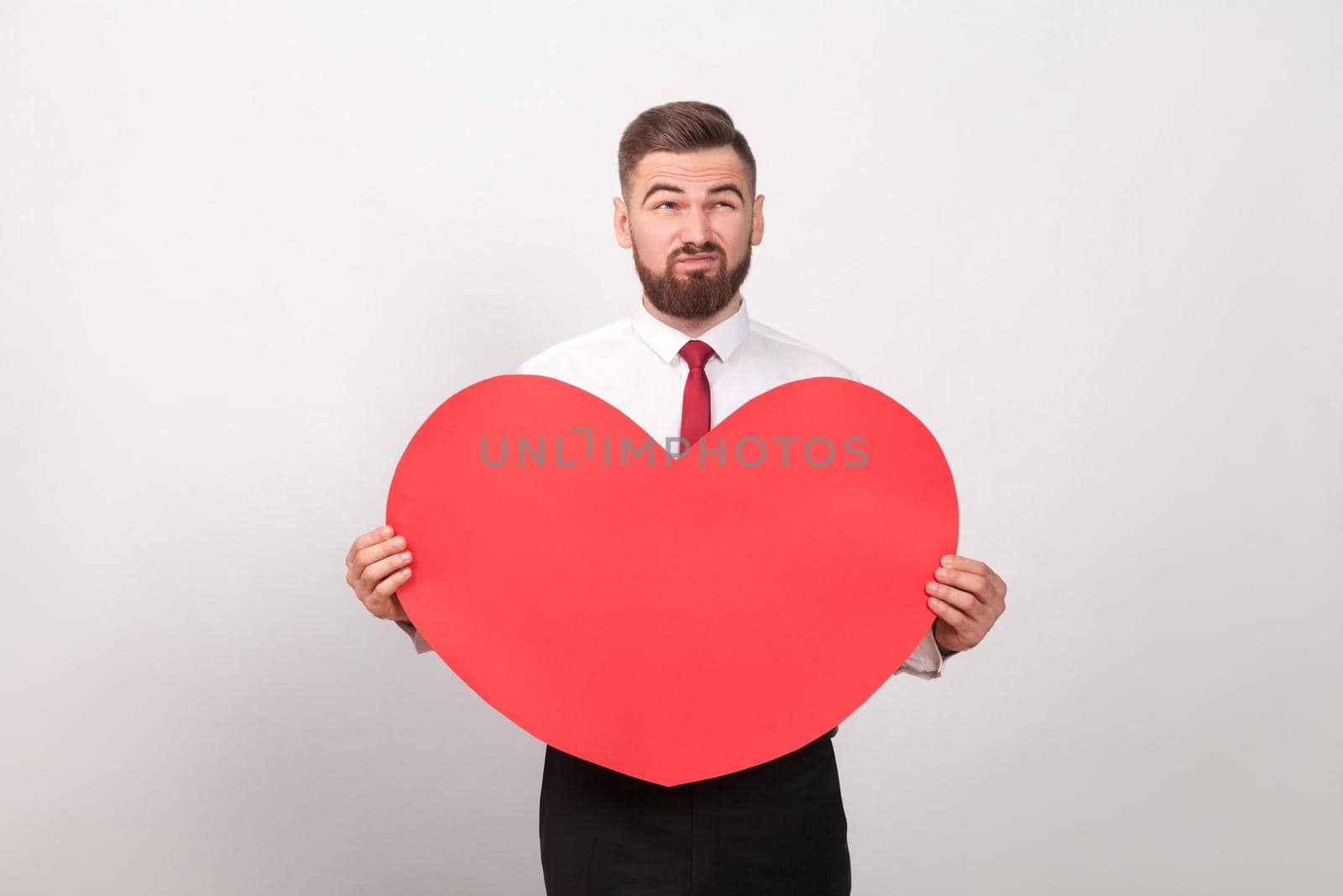 Bearded man thoughtful, holding red heart and thinking of love. Indoor, studio shot, isolated on gray background